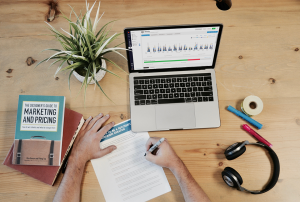 a hand writes on a piece of paper in front of a laptop and a book on a table