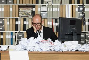a man sits at a desk with a pile of papers and a dell monitor