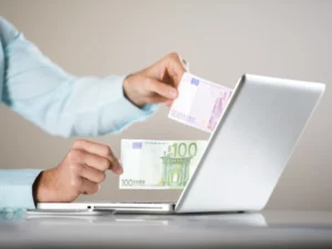 en person, der holder en 100 euro-seddel foran en bærbar computer