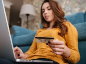 a woman is sitting on a couch holding a credit card and using a laptop computer .