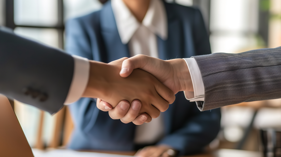 två kostymklädda personer skakar hand medan en dam står framför dem