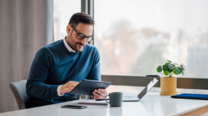 a guy on glasses looks at his tablet with a cup and a laptop on the table in front